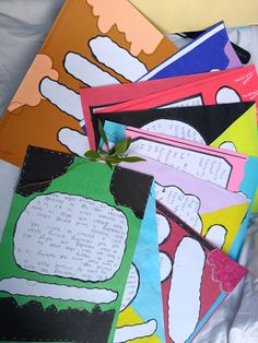 several colorful papers with writing on them sitting on a white sheeted surface, one is folded in half and the other has a green plant growing out of it's leaves