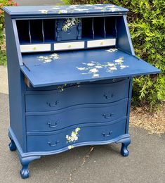 a blue desk with flowers painted on the top and bottom drawer, sitting in front of some bushes