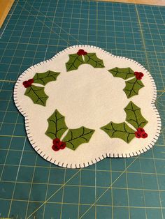 a piece of felt with holly leaves and red berries is sitting on a cutting board