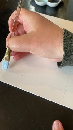 a hand holding a paintbrush over a piece of paper on top of a table