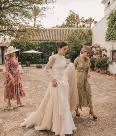 three women in dresses and hats are standing on the dirt ground near an outdoor area