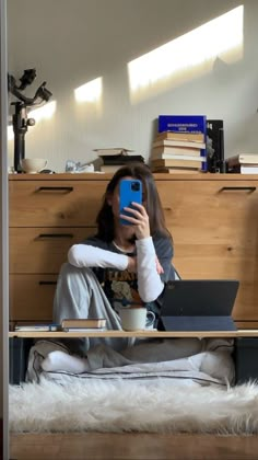 a woman sitting on the floor with her laptop and holding a cell phone in front of her face