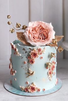 a blue cake with pink flowers on top and gold leaves around the edges, sitting on a white table