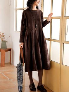 a woman standing next to a window wearing a brown dress and holding a handbag