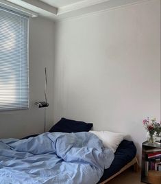 an unmade bed with blue sheets in a white room next to a book shelf