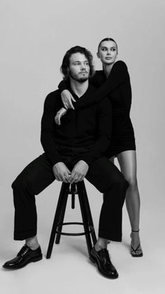 two people sitting on top of a stool posing for a black and white photo with their arms around each other