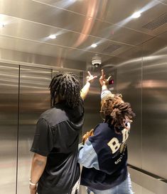 two people standing in front of an elevator taking pictures with their cell phones on the mirror