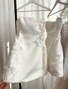 two wedding dresses hanging on a rack in front of a window with white drapes