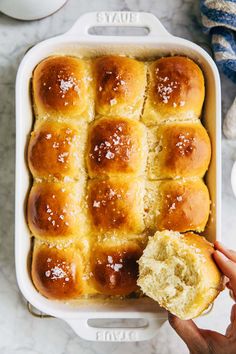 a person holding a piece of bread in front of a casserole dish filled with rolls