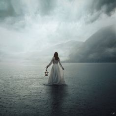 a woman in a long white dress holding a lantern standing on the edge of a body of water