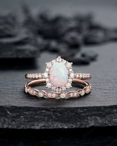 a white opal and diamond ring sitting on top of a black stone slab with flowers in the background