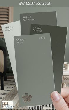 a hand holding three different shades of gray paint in front of a living room chair