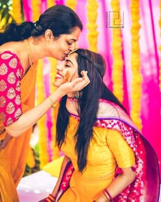 two women in yellow and pink saris touching each other's cheek while smiling