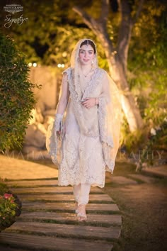 a woman in a white dress and veil walking down the street with trees behind her