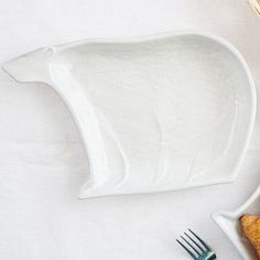 a white bear shaped dish next to a fork and bowl with bread on the side