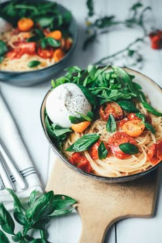 two bowls filled with pasta and vegetables on top of a wooden cutting board next to silverware