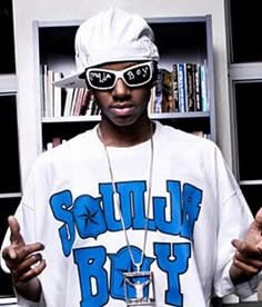 a man wearing sunglasses and a white shirt with blue writing on it, standing in front of a bookshelf