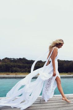 a woman in white dress walking on wooden pier next to blue water with trees in the background