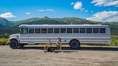 two people sitting in front of a bus on a dirt road with mountains in the background