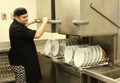 a man working in a kitchen with lots of white plates on the rack and silver dishwasher