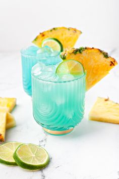 two glasses filled with blue liquid next to sliced pineapples and lime wedges