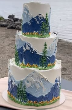 a three tiered cake decorated with mountains and pine trees on a table near the water