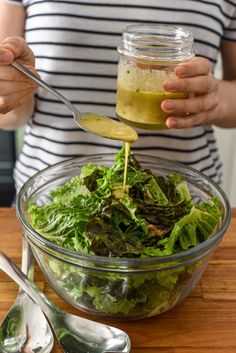 a person pouring dressing into a salad in a glass bowl on top of a wooden table