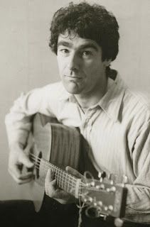 a black and white photo of a man holding a guitar in his right hand while looking at the camera