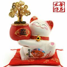 a white cat figurine sitting on top of a red pillow next to a tree