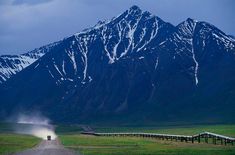 a truck driving down a dirt road in front of snow covered mountains and green grass