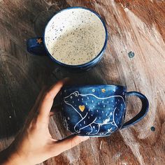 a hand holding a blue mug on top of a wooden table next to another cup