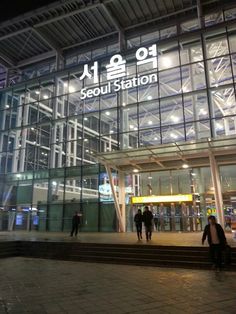 people are walking in front of the seoul station building at night, with lights on