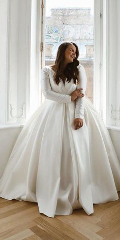 a woman standing in front of a window wearing a long white dress and holding her hands on her hips