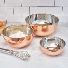 three metal bowls and a whisk on a white marble countertop next to a cutting board