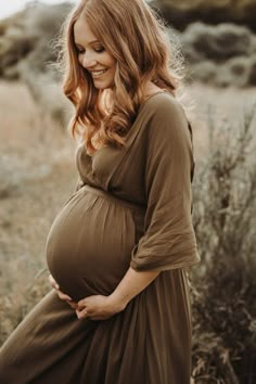 a pregnant woman in a brown dress is standing in a field and smiling at the camera