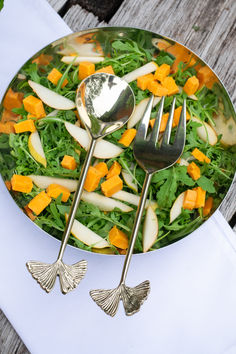 a salad with apples, carrots and spinach on a plate next to two silver spoons