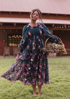 a woman standing in the grass holding a basket and wearing a dress with flowers on it