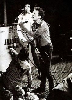 black and white photograph of two men on stage with microphones in their hands, surrounded by other people