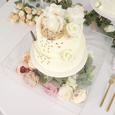 a wedding cake with white flowers and greenery on the table next to gold utensils