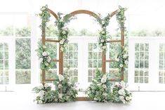 an arch decorated with flowers and greenery stands in front of two large windows that look out onto the outdoors