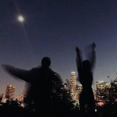 two people standing in front of a city skyline at night with their arms raised up