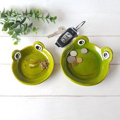 two green frog shaped dishes with keys in each one's eyes, on a white wooden surface