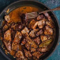 a pan filled with meat and sauce on top of a blue countertop next to a wooden spoon