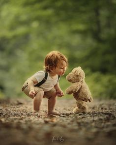 a little boy playing with a teddy bear in the middle of a dirt road surrounded by trees