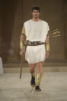 a man in an ancient greek costume walks down the runway with his hands on his hips