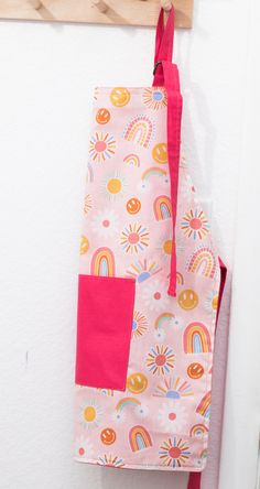 a pink apron hanging on a wall with a wooden hanger and rainbows printed on it