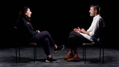 two people sitting on chairs facing each other in the dark, one is talking to the other