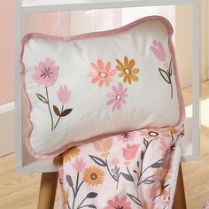 a white bed with pink and yellow flowers on it, next to a wooden chair