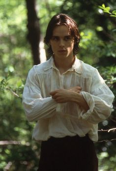 a man standing in the woods wearing a white shirt and black pants with his arms crossed