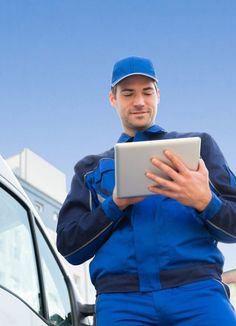 a man in blue overalls holding a tablet next to a white van and looking at the camera